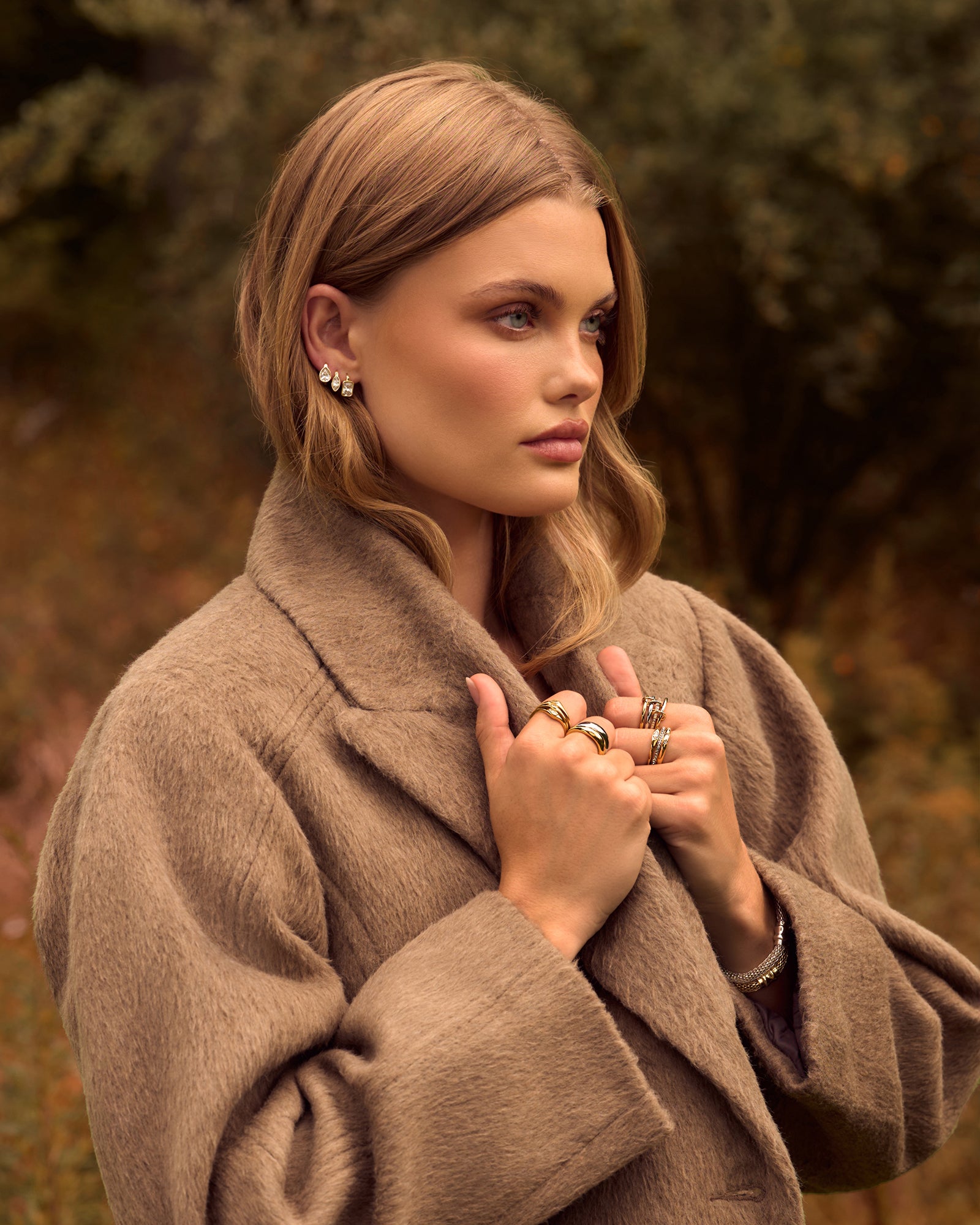 Model in a camel coat outdoors wearing a gold crystal ear stack, stacked rings and a bracelet
